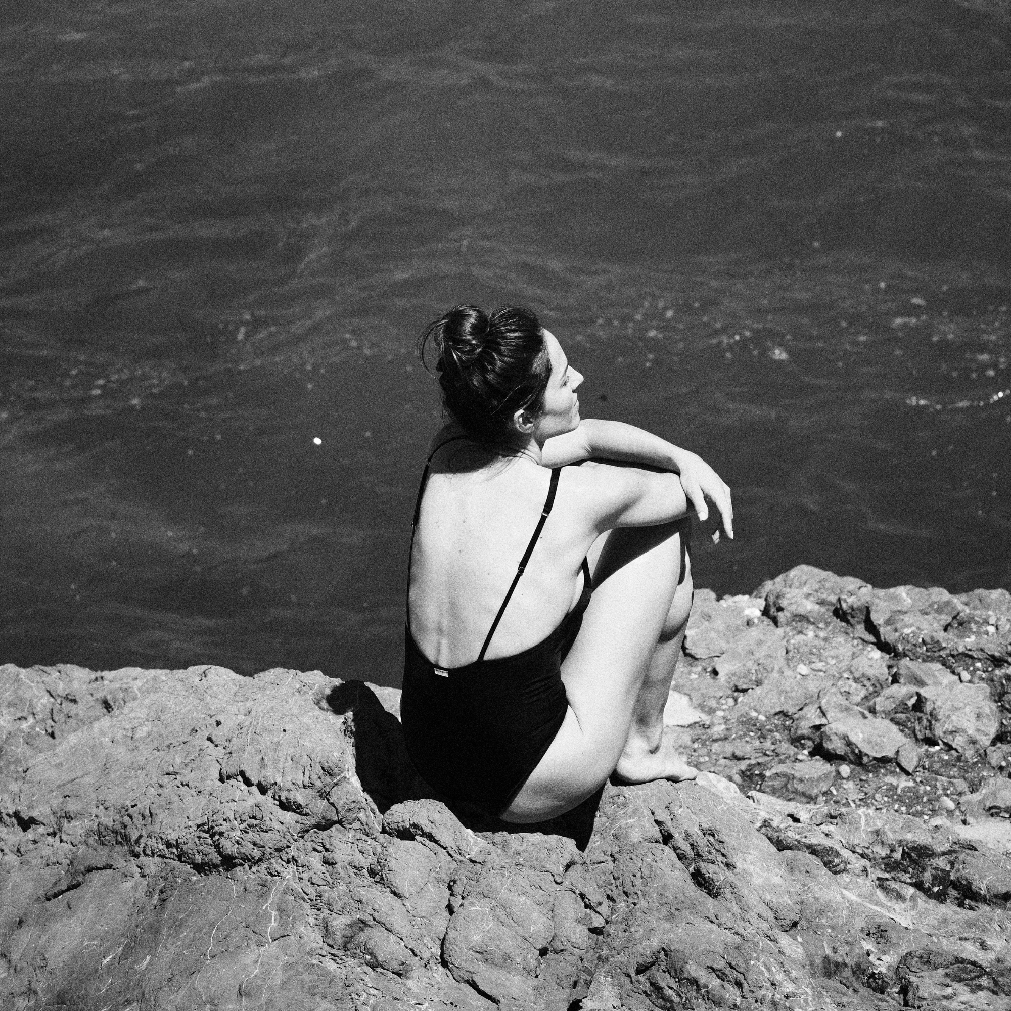 Woman in a black swimsuit by Paupa sitting on rocks by the sea